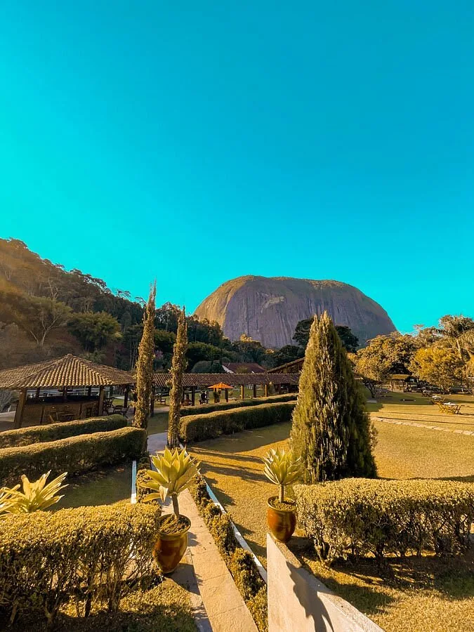 Paisagem da fazenda com construção amarela e a Pedra do Rosário ao fundo